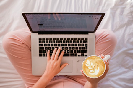 Jeune femme assise sur son lit consulte son portable avec une tasse de café